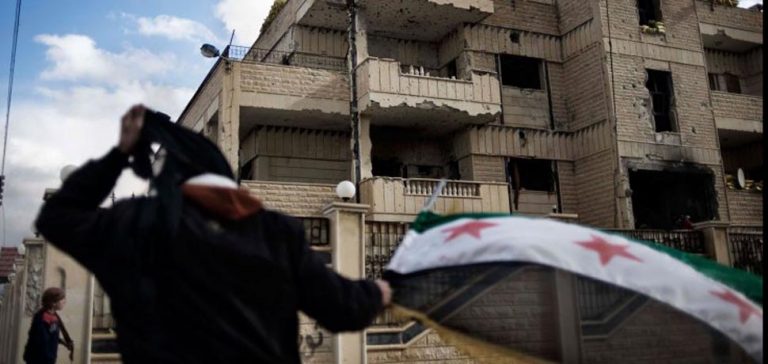 A man waves the old Syrian flag in front of the former police st, cc Flickr Freedom House, modified, https://creativecommons.org/licenses/by/2.0/
