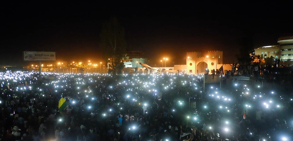 On 7 April 2019, the second day of the sit-in, protestors were gathering infront of the Military Headquarter chanting for freedom, piece, and justice. People in the crowd told each other to turn on their phone torches., cc Ola A .Alsheikh, modified, https://commons.wikimedia.org/w/index.php?search=khartoum+protest+filetype%3Abitmap&title=Special%3ASearch&profile=advanced&fulltext=1&advancedSearch-current=%7B%22fields%22%3A%7B%22filetype%22%3A%22bitmap%22%7D%7D&ns0=1&ns6=1&ns14=1#/media/File:Sudan_sit-in.jpg