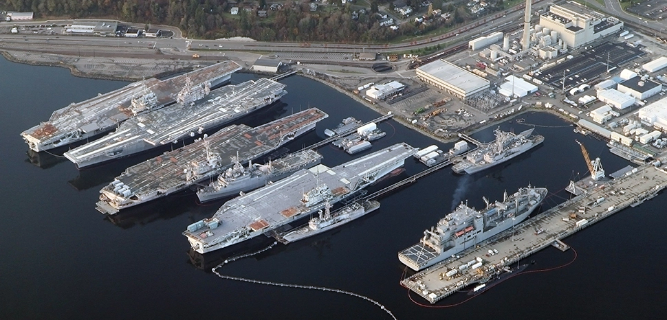 cc Jelson25, modified, An aerial view of the Puget Sound Naval Shipyard and Intermediate Maintenance Facility in Bremerton, Washington (USA), on 24 November 2012. Visible are the following ships (top to bottom): aircraft carrier USS Independence (CV-62); USS Kitty Hawk (CV-63); USS Constellation (CV-64); amphibious transport dock ship USS Dubuque (LPD-8); USS Ranger (CV-61); three Oliver Hazard Perry-class frigates; an (active) Lewis and Clark-class dry cargo ship; a Seawolf-class submarine. - https://commons.wikimedia.org/wiki/File:Aerial_Bremerton_Shipyard_November_2012.jpg
